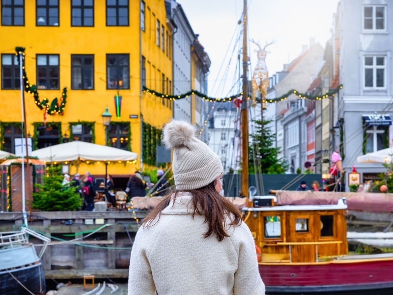 Woman in a cream bobble hat facing boats in a canal and colorful buildings
