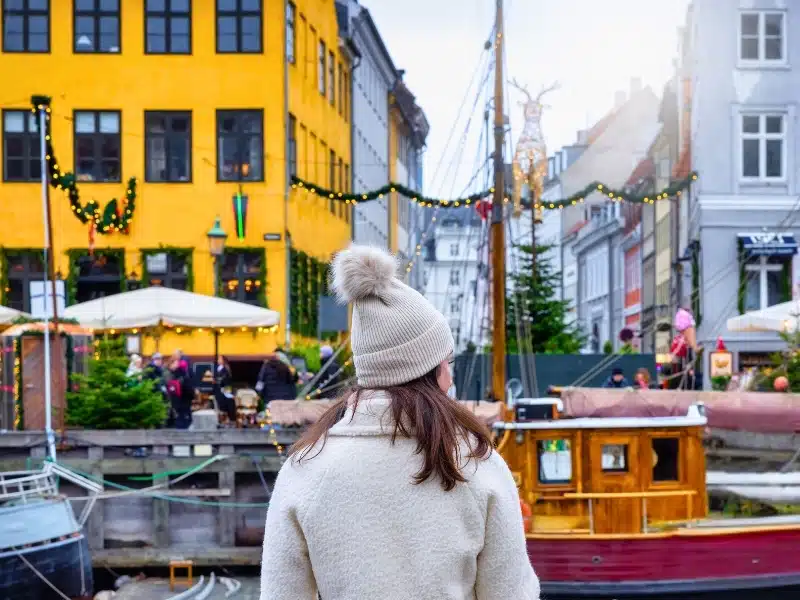 Woman in a cream bobble hat facing boats in a canal and colorful buildings