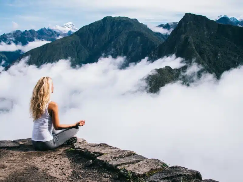 Back view of woman tourist sitting in lotus pose relaxing and admiring breathtaking view during trek tour, female traveler enjoying beauty of Machu Picchu landscape and high mountains covered clouds