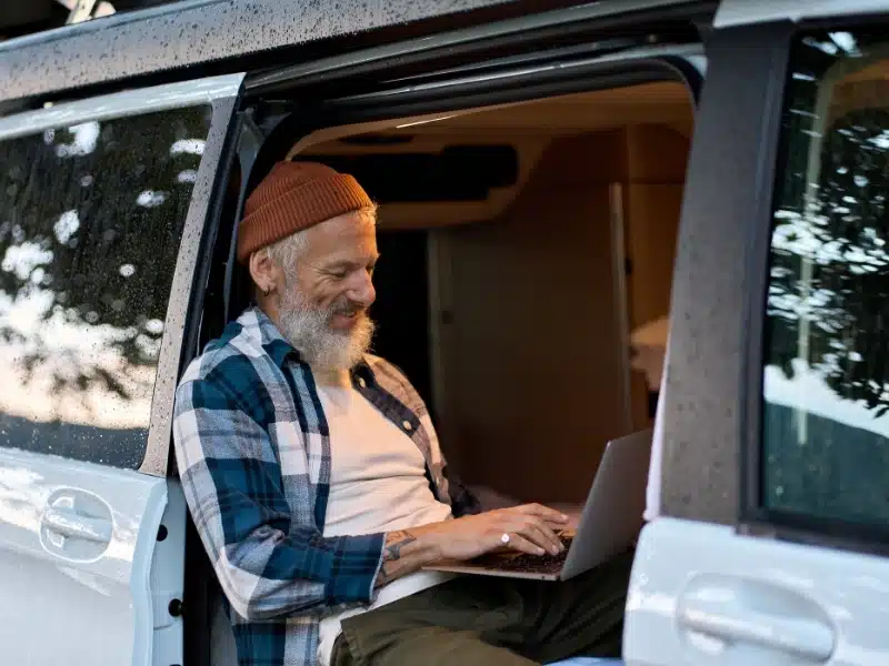 Happy older man sitting in rv camper van using laptop. Smiling mature active traveler holding computer on lap remote working online and enjoying vanlife, freedom, resting in outdoor camping.