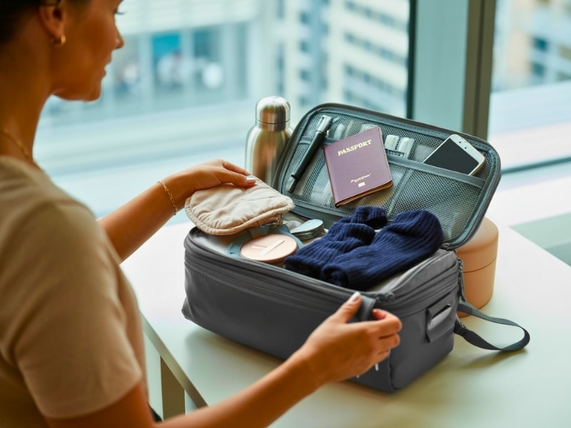 A photograph taken over the shoulder of a woman carefully packing a compact, dark grey travel bag.