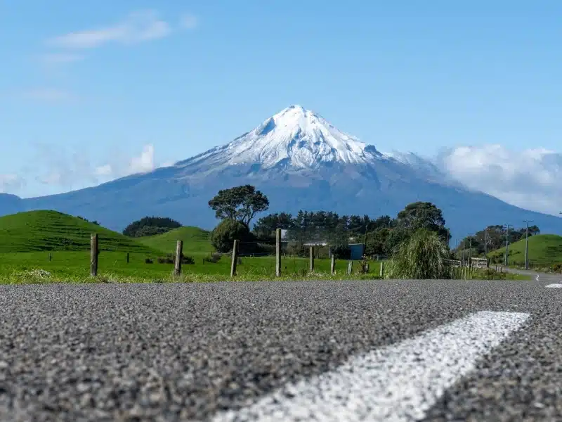 A scenic road leads to the snow-capped Mount Taranaki under a clear blue sky in New Zealand's lush countryside.