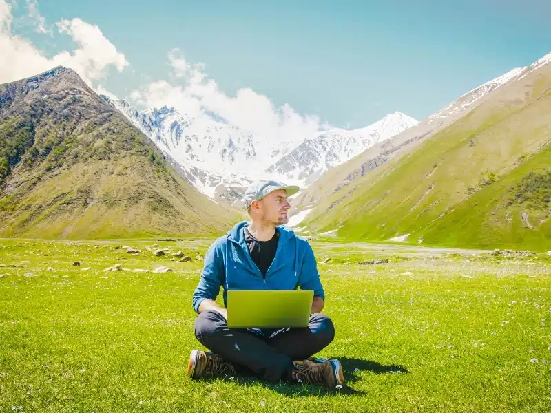 Freelancer digital nomad sits work online with mountains, meadows and scenic panorama surrounding him