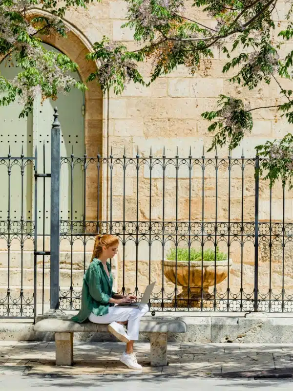 redhead woman sitting on concrete bench and using laptop near forged fence in Valencia, Spain