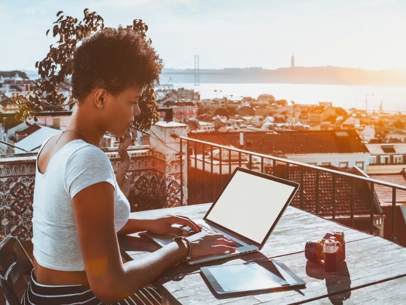 Young appealing biracial female photographer sitting on balcony of Lisbon hotel with stunning bright cityscape in background and working with laptop mock-up; retro photo camera and tablet pc near her