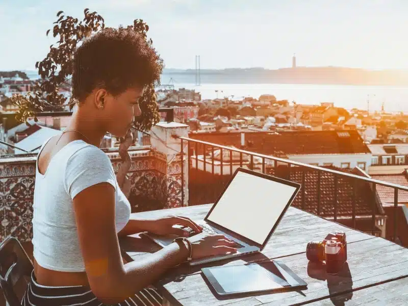 Young appealing biracial female photographer sitting on balcony of Lisbon hotel with stunning bright cityscape in background and working with laptop mock-up; retro photo camera and tablet pc near her