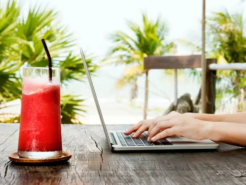 Woman using a laptop on a wooden table with a red drink by the beach