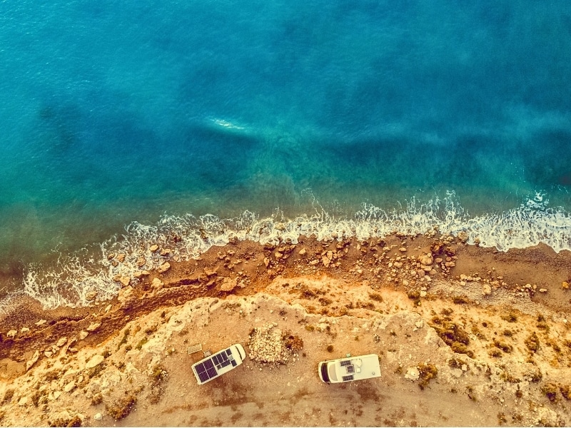 Aerial view. Two camper vehicles with solar photovoltaic panels on roof camping on sea, coastal cliff in Spain. 