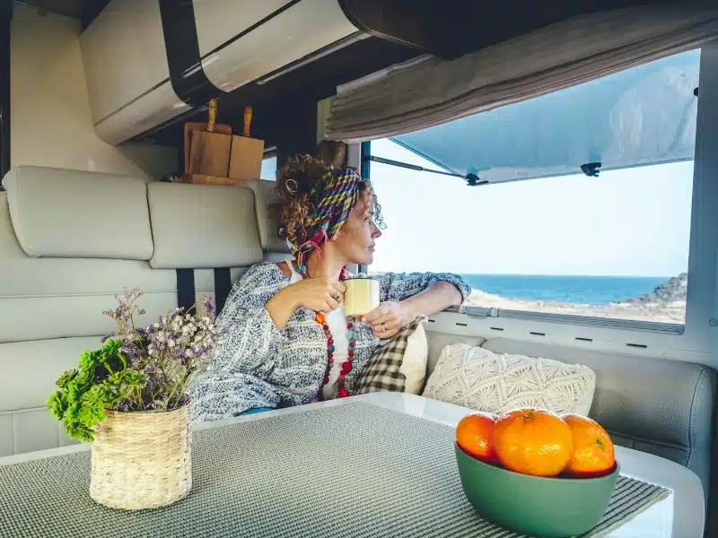 Adult woman drinking and enjoying sea view inside camper van motorhome comfortably on the sofa. 