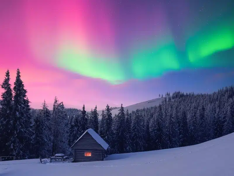 Small wooden cabin in snowy mountains of Norway under incredible Northern lights in starry sky. 