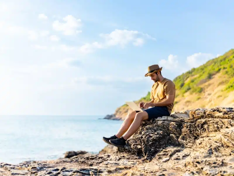 Man sitting on a rocky coastline and working on laptop computer 