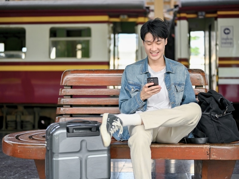 Male traveler using his smartphone while waiting for his train on a bench at the platform.