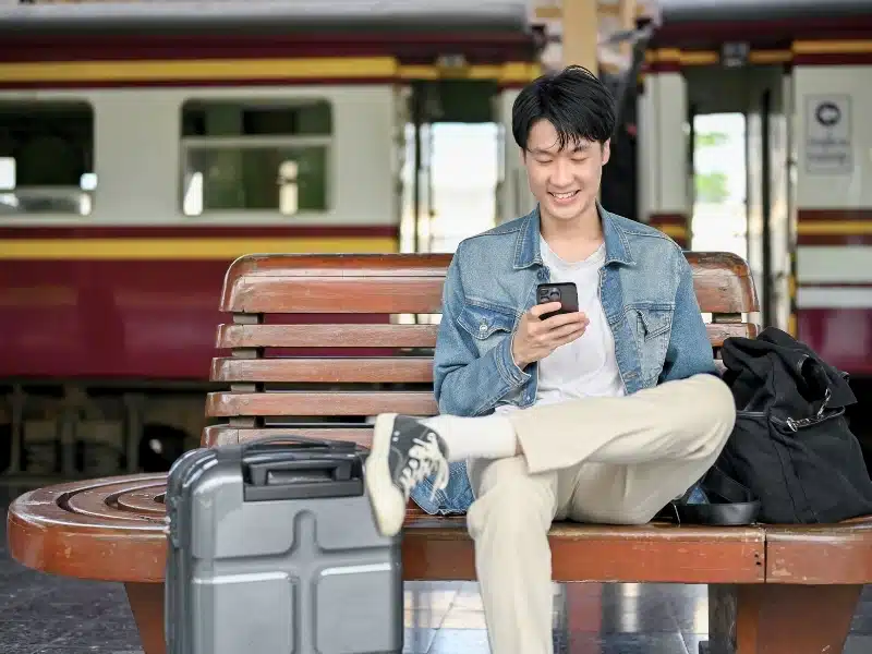 Male traveler using his smartphone while waiting for his train on a bench at the platform.