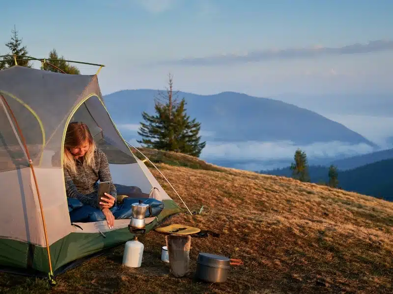 Woman traveler sitting in camp tent and taking photo of tourist gas burner and kettle. Female hiker using mobile phone while resting in tourist tent on grassy hill with mountain and sky on background.