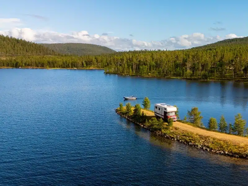 A cozy camper sits on a lush green peninsula facing a serene blue lake surrounded by dense forest. 