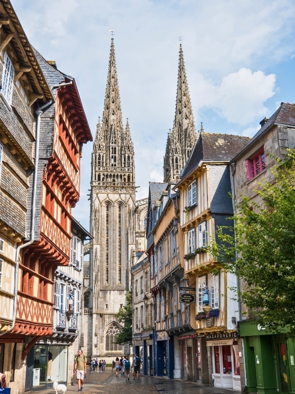 Half timbered houses line a cobbled street with a Gothic cathedral in the background