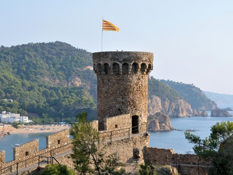 Detail of one round stone tower of a walled city above the sea, flying a yellow and red striped flag