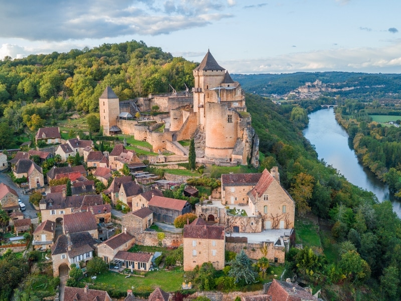 creamy stone turreted castle and village overlooking a river lined with trees