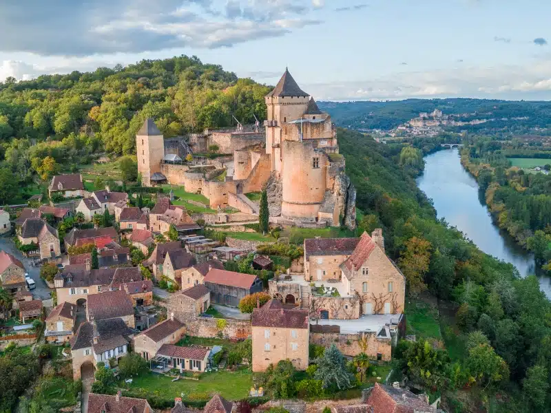 creamy stone turreted castle and village overlooking a river lined with trees