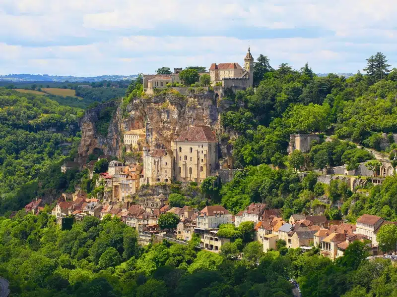 Small village of stone houses and red roofs with a tall cliff behind topped with a church and castle