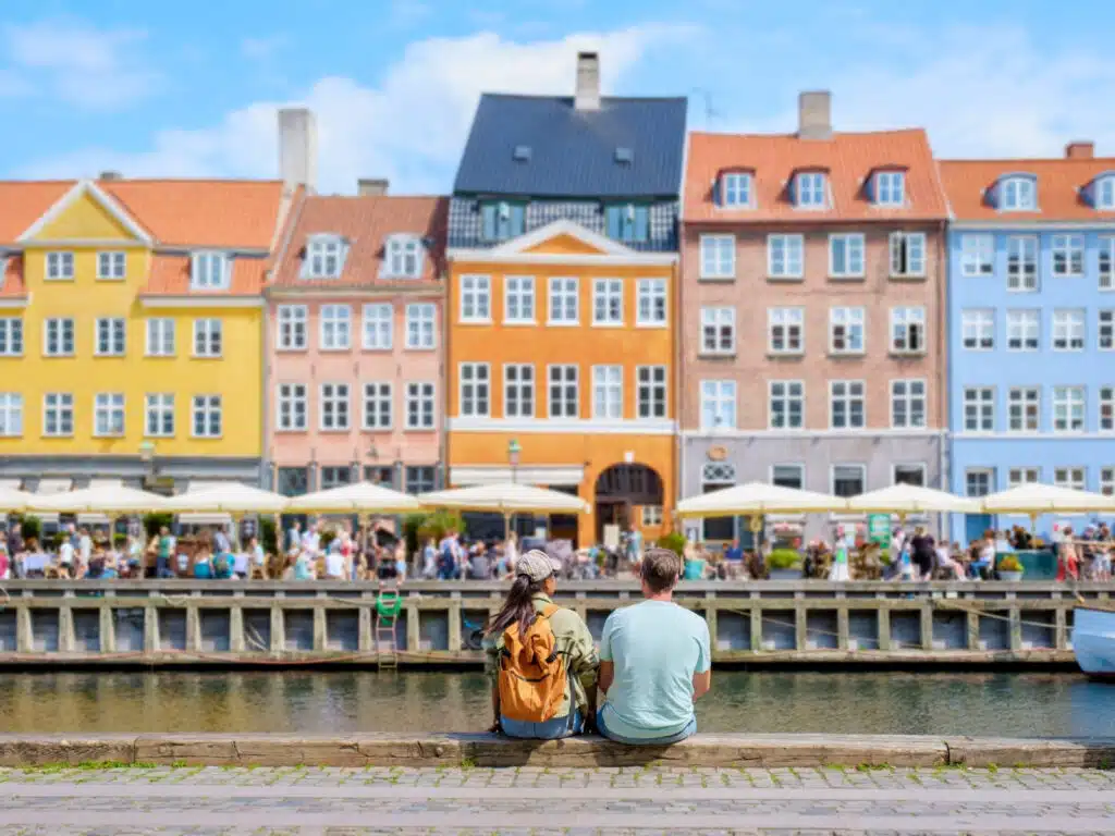 A couple sits on a stone wall overlooking a bustling Copenhagen canal, taking in the vibrant facade of historic buildings and the lively atmosphere of the waterfront.