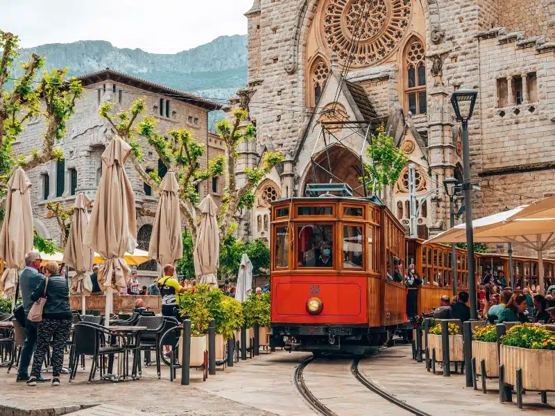 Mallorca, Spain. April 27, 2022. Orange tram moving on railroad tracks against church and amidst restaurants. Male and female tourists exploring city. Church of Sant Bartomeu de Soller.