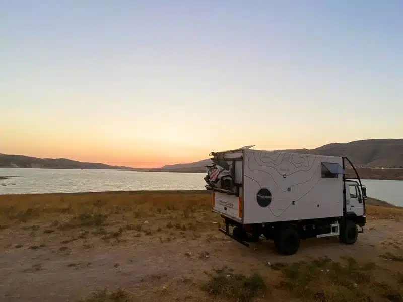 overland truck parking by a large body of water at sunset