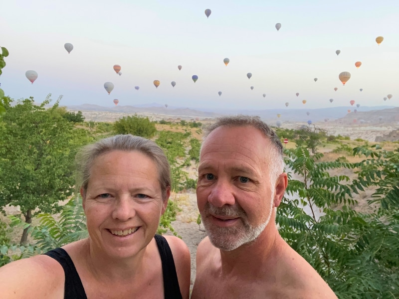 couple standing on front of many hot air balloons and surrounded by greenery