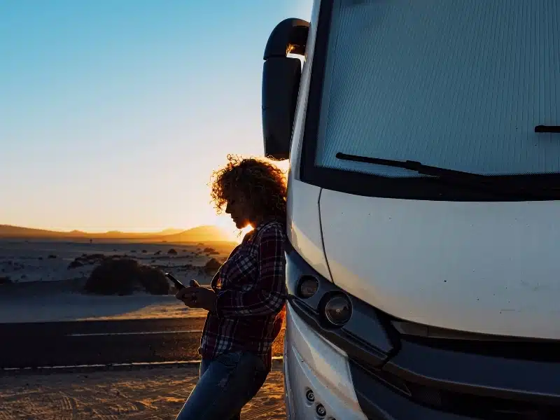 People on travel with modern camper. Woman using mobile phone roaming esim against big motorhome.