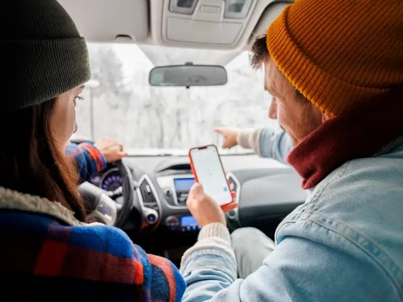 Cheerful couple having fun driving a car while travelling to a winter vacation road trip, using smart phone with esim