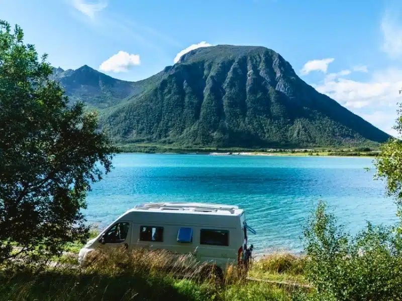 Discover Lofoten, Norway with a camper van parked by a breathtaking lake. Towering mountains and lush greenery surround the tranquil waters under a clear blue sky.