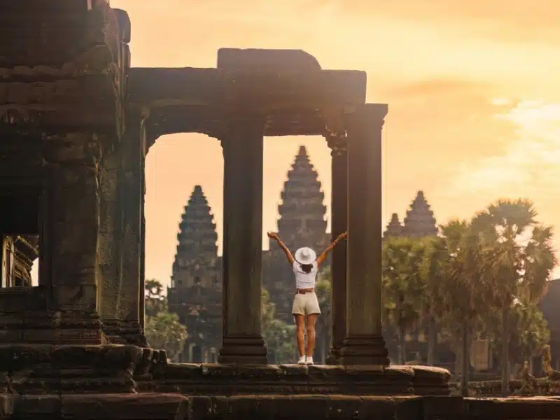 Female backpacker in white hat stretches her arms between stone columns at Angkor Wat, sunrise glow over palms and towers. Concept, high quality travel image for tourism ads, blogs and guides.