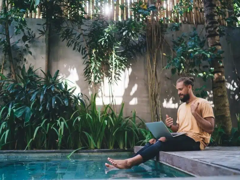 Side view of bearded male in casual clothes sitting on edge of swimming pool with netbook and communicate on video chat. Digital nomad working online on freelance