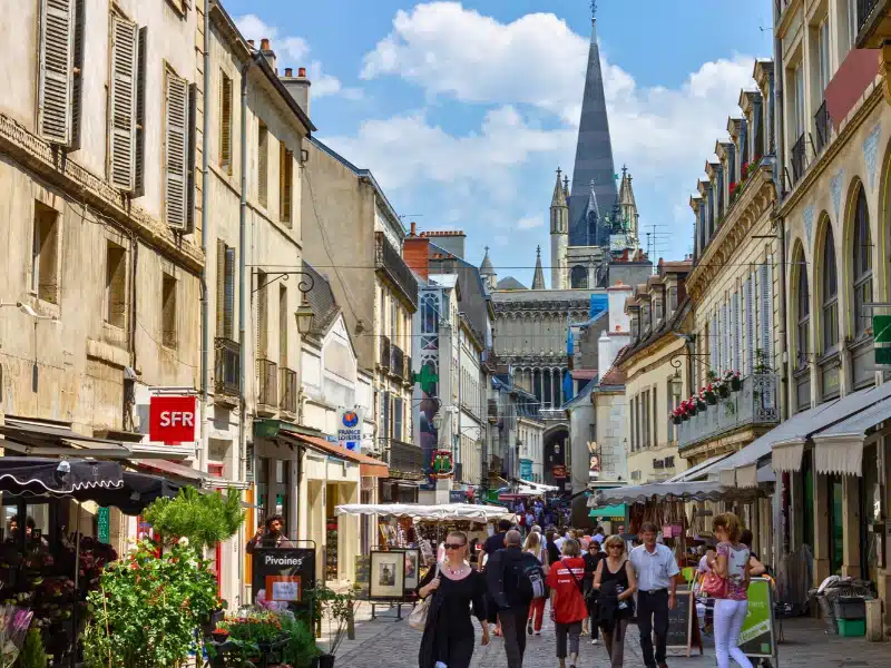 The historical buildings and restaurants with tourists and Notre Dame church tower against blue cloudy sky on a sunny day, Dijon, France