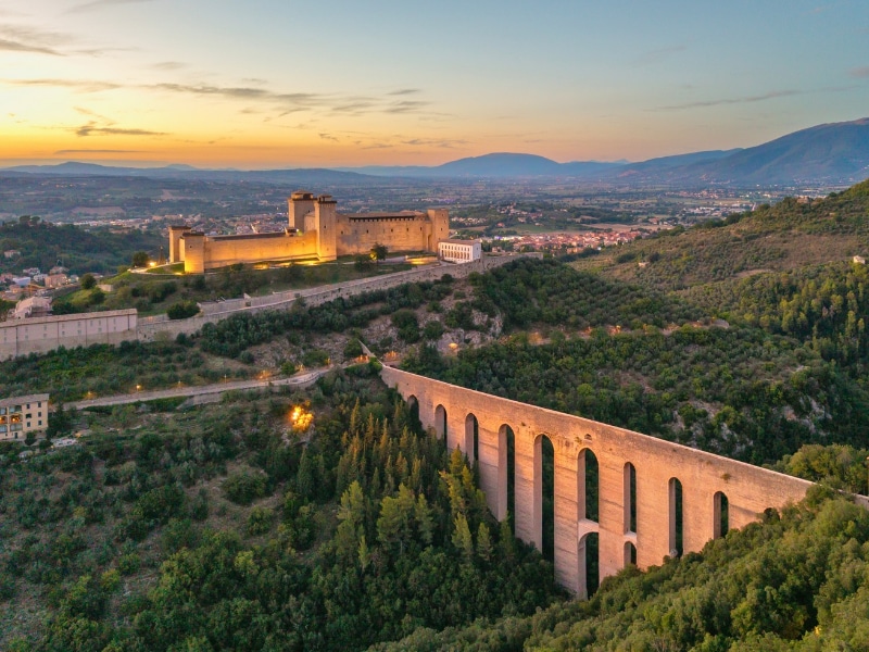 Dramatic aerial view of medieval Spoleto, Umbria at twilight. Features the illuminated Ponte delle Torri aqueduct and Rocca Albornoziana fortress glowing under city lights in Italy.