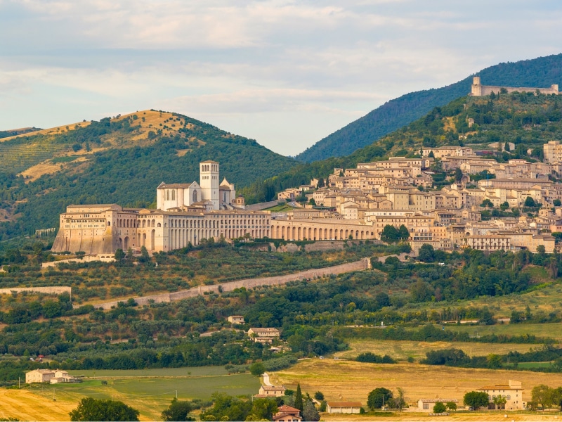 Aerial view of the Basilica of Saint Francis, Assisi, Italy, showcasing Romanesque and Gothic architecture. The iconic white and stone structure contrasts against lush hills under clear daylight.
