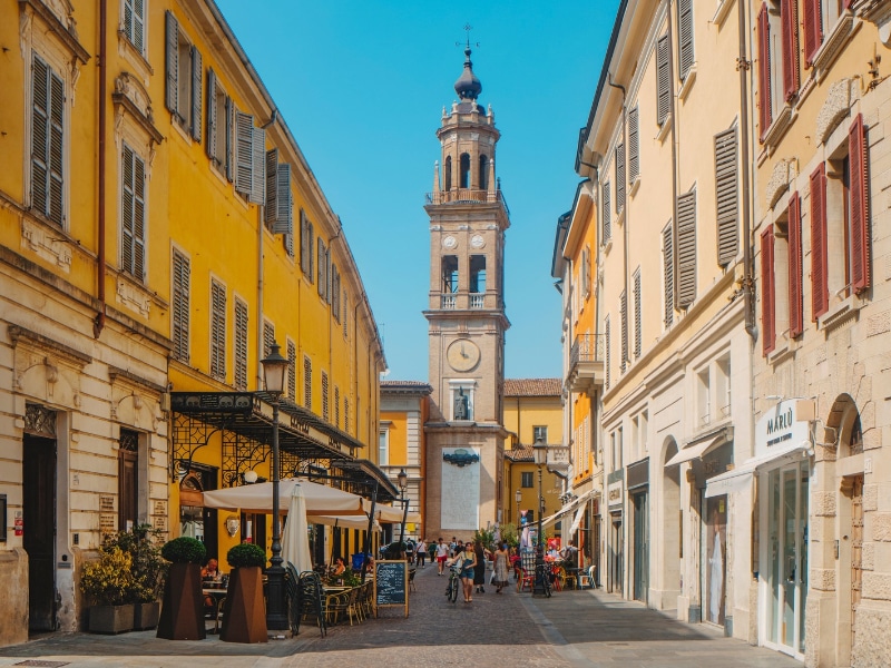 Parma, Italy: A view along Strada Cavour, with lively cafes, shoppers and cyclists framed by yellow facades, leading toward the distant Monumento ai Caduti in the background
