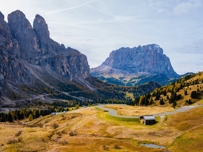 In the breathtaking Dolomites of Italy, early autumn casts a golden hue on the rugged mountains, revealing winding roads and a charming cabin nestled in the serene valley.
