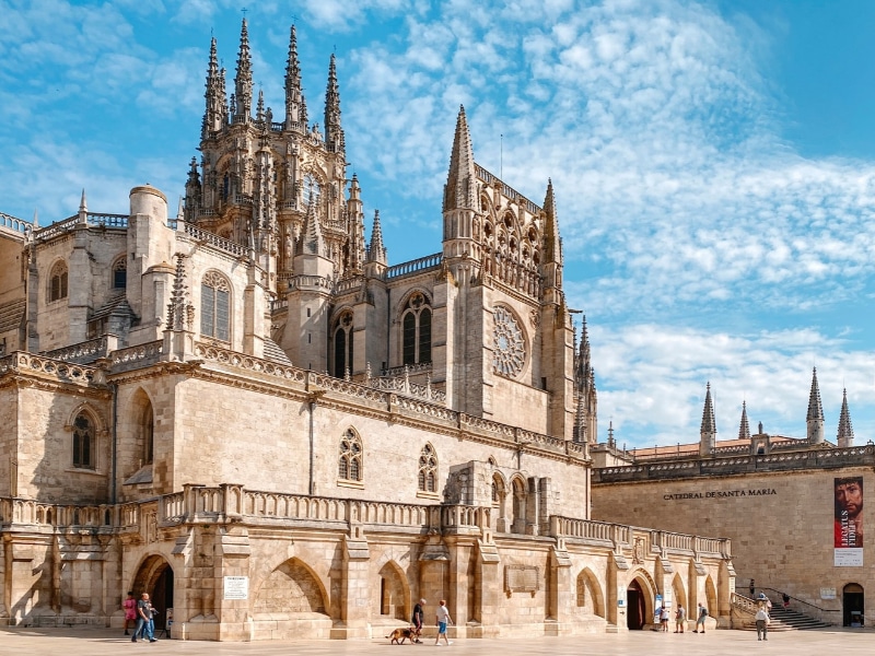 Visitors approaching the entrance of the impressive Cathedral of Burgos from Plaza del Rey San Fernando, under a clear blue sky