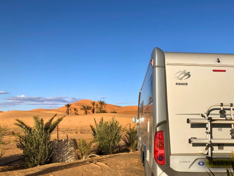 silver motorhome parked by large sand dunes and surrouned by palm trees