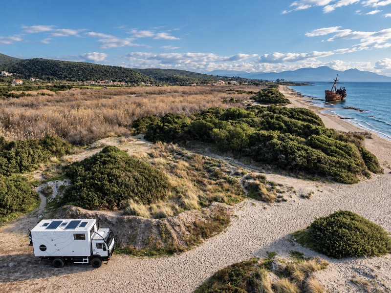 Overland truck parked among coastal sand dunes overlooking a rusting shipwreck on the beach in southern Greece, with the sea and mountains beyond.