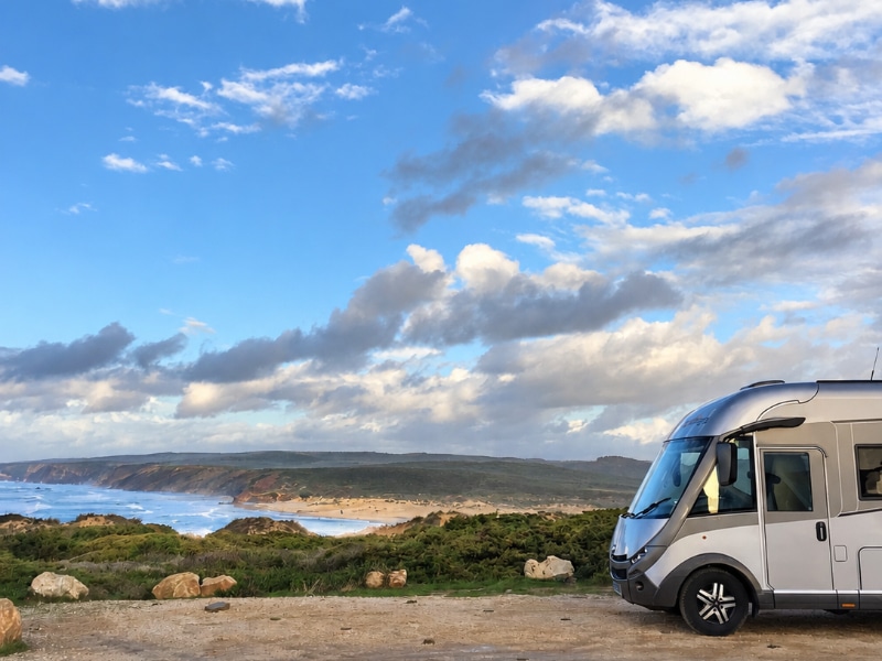 Motorhome parked above a sandy beach, with coastal cliffs and a blue sky dotted with clouds.